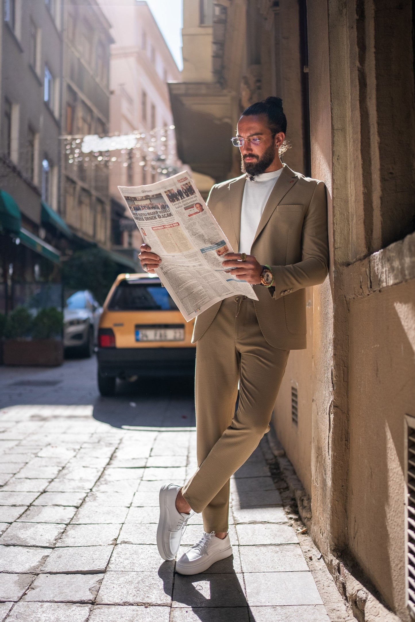 Man in a beige suit reading a newspaper on a city street.