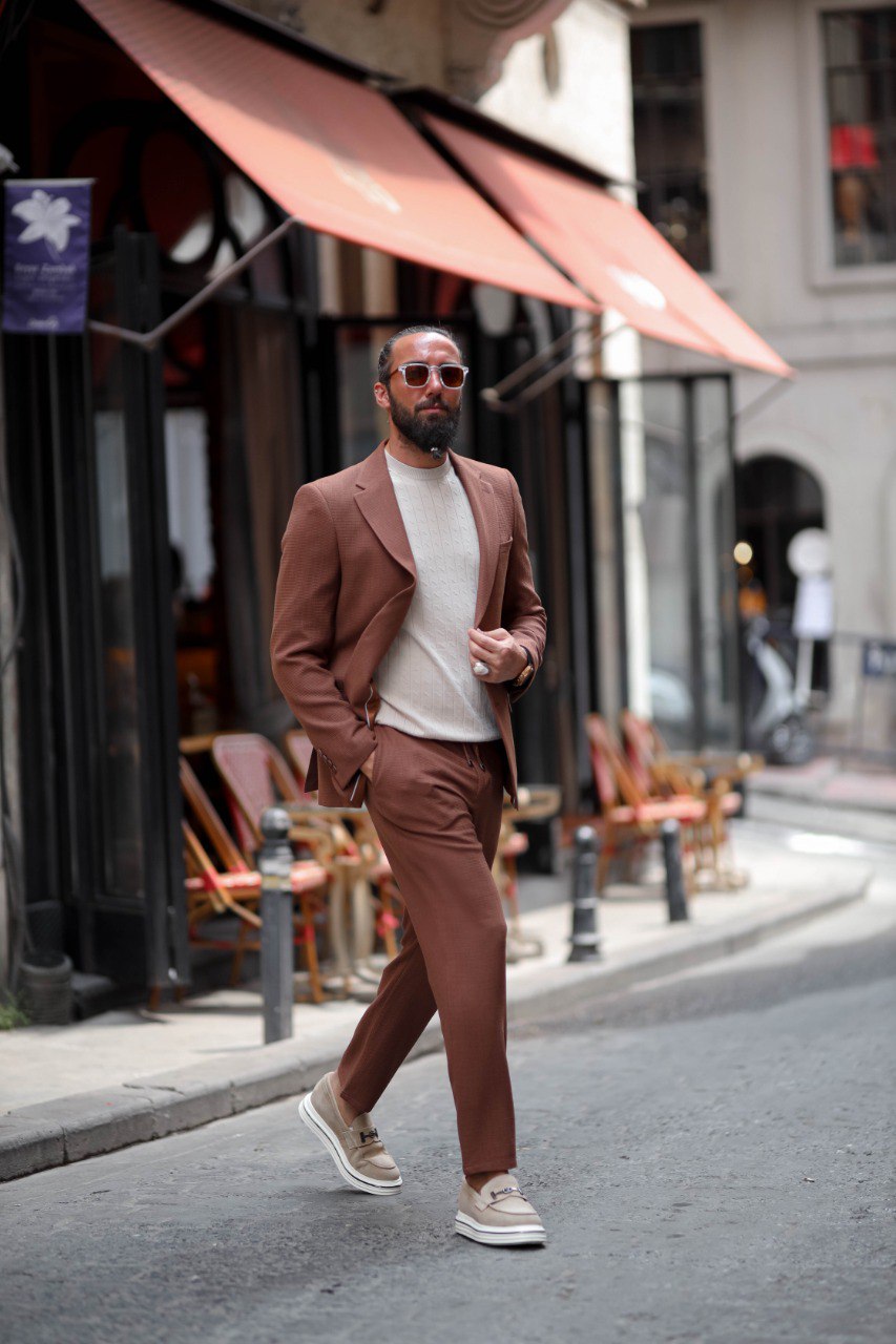 Man in a brown suit walking on a city street with awnings and chairs in the background.