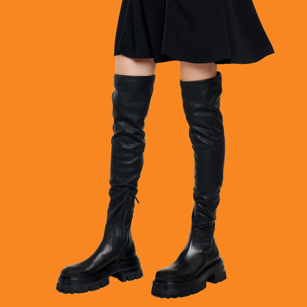 Black knee-high boots on a person wearing a black skirt against an orange background