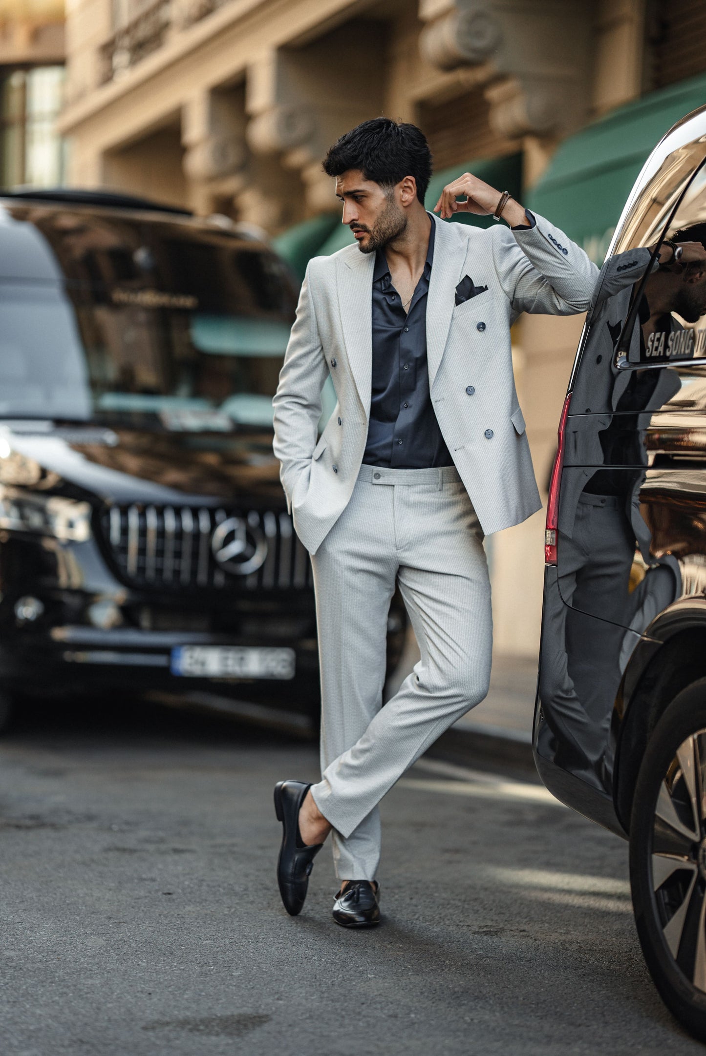 Man in a light gray suit standing next to a luxury car in an urban setting