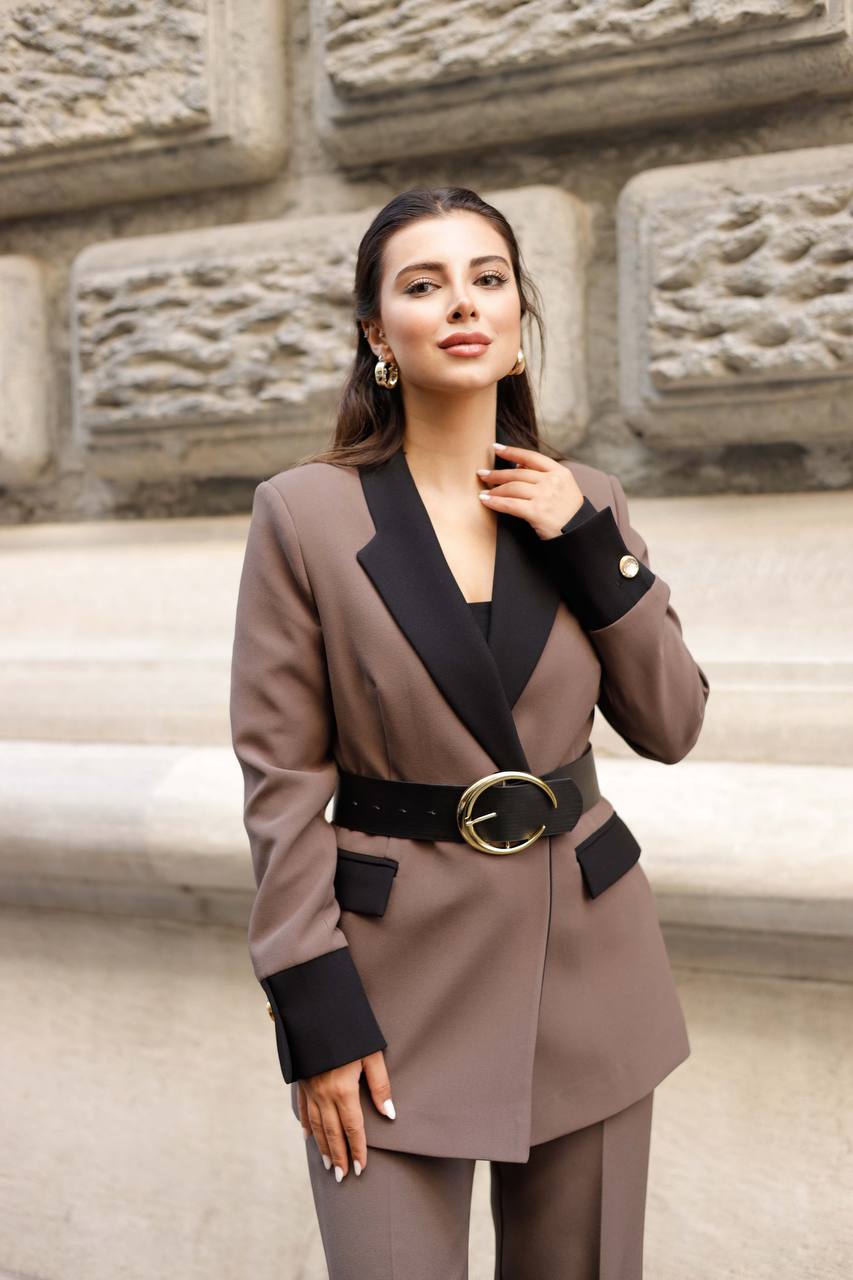 Woman wearing a stylish brown and black suit with a gold belt against a stone wall.
