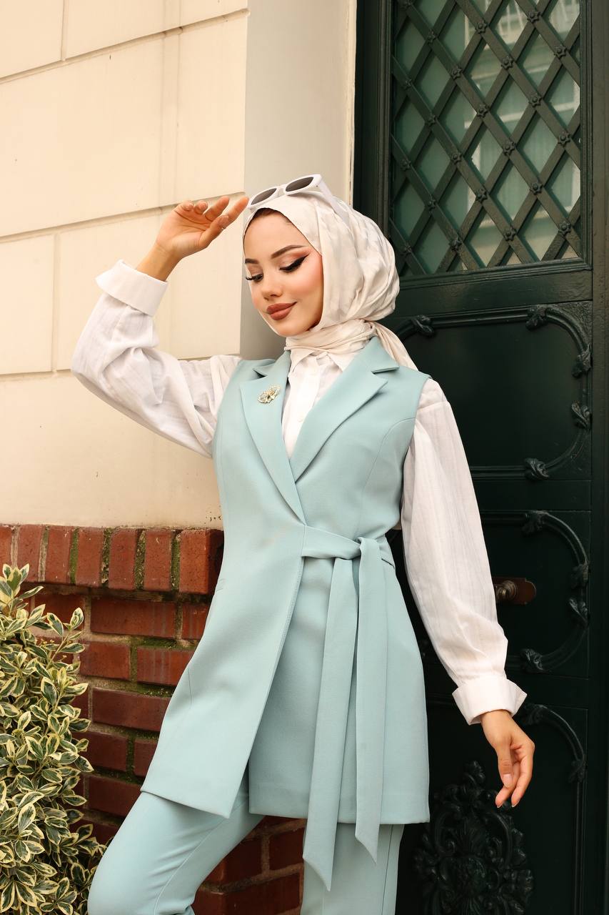 Woman wearing a light blue vest over a white shirt, standing in front of a door.