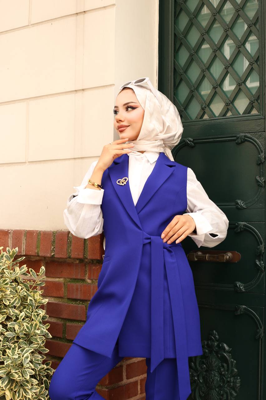 Woman wearing a blue vest and white hijab standing in front of a green door.