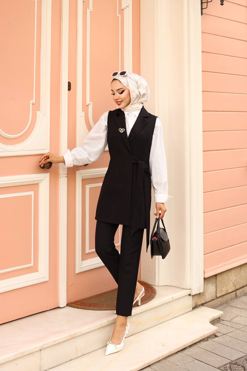 Woman in a black vest and pants outfit standing in front of a pink door.