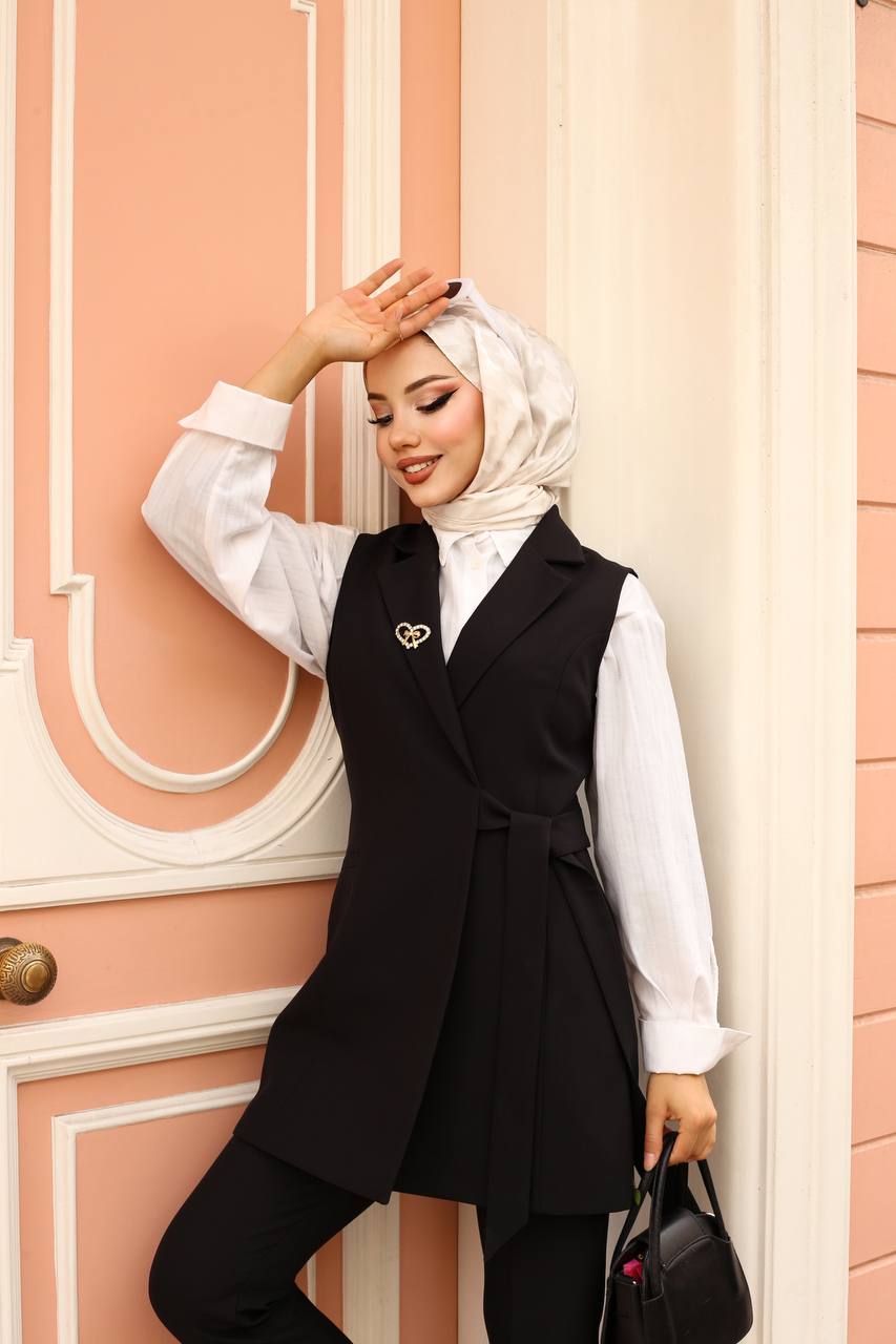 Woman wearing a black vest over a white shirt with a hijab, standing against a pink wall.