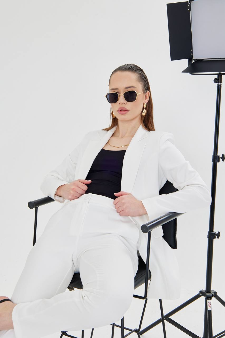 Person wearing a white blazer and black top sitting on a chair with studio lighting equipment in the background.