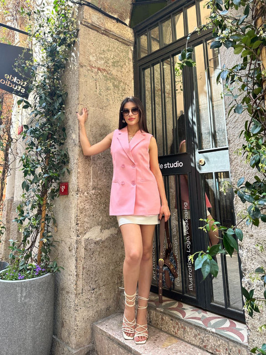 Woman in a pink vest and white shorts standing against a building with plants and a store entrance.