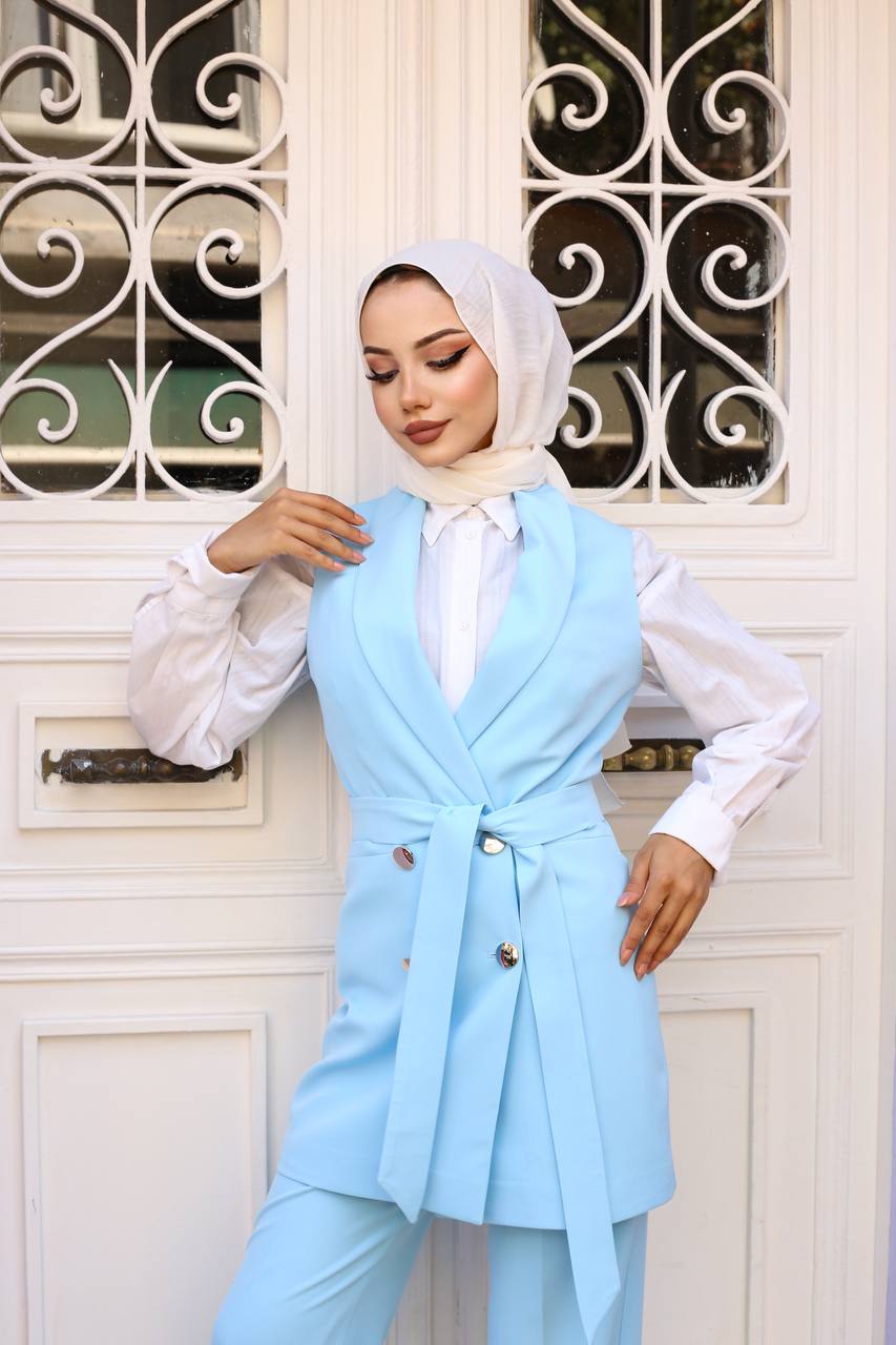Woman wearing a light blue blazer over a white shirt with a white hijab, standing in front of a decorative metal gate.