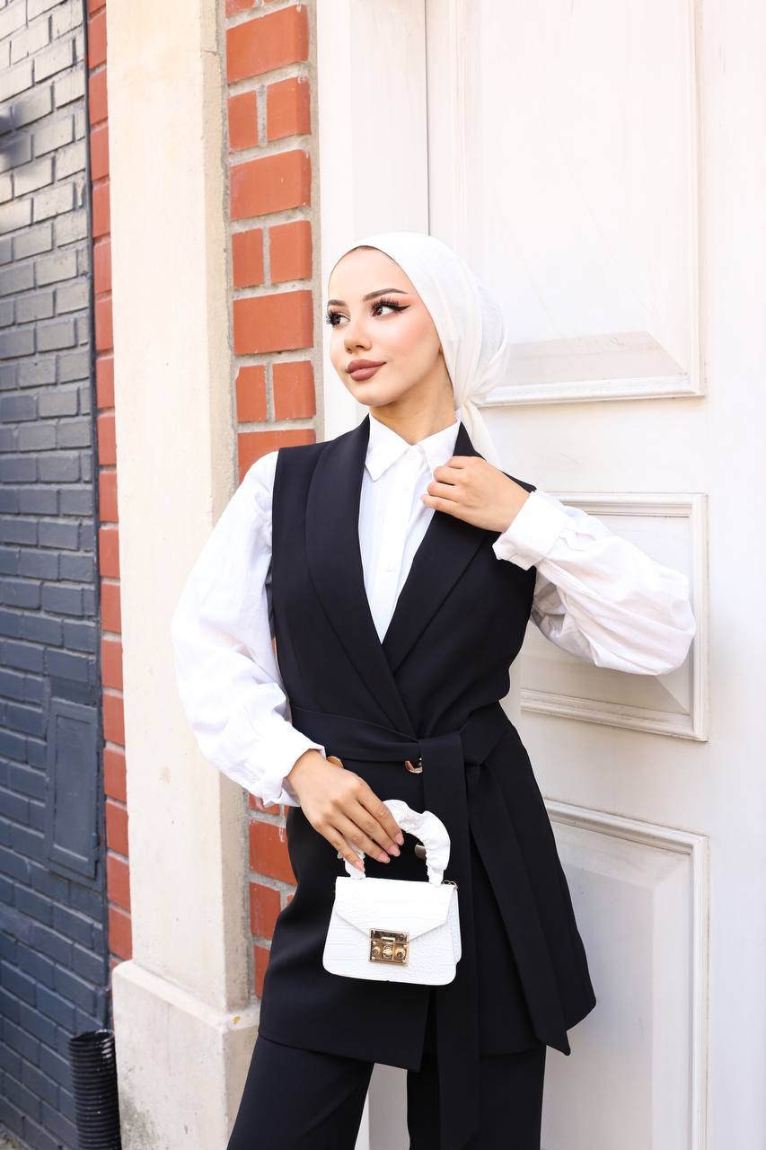 Woman in a black vest and white shirt holding a small white handbag against a brick wall.