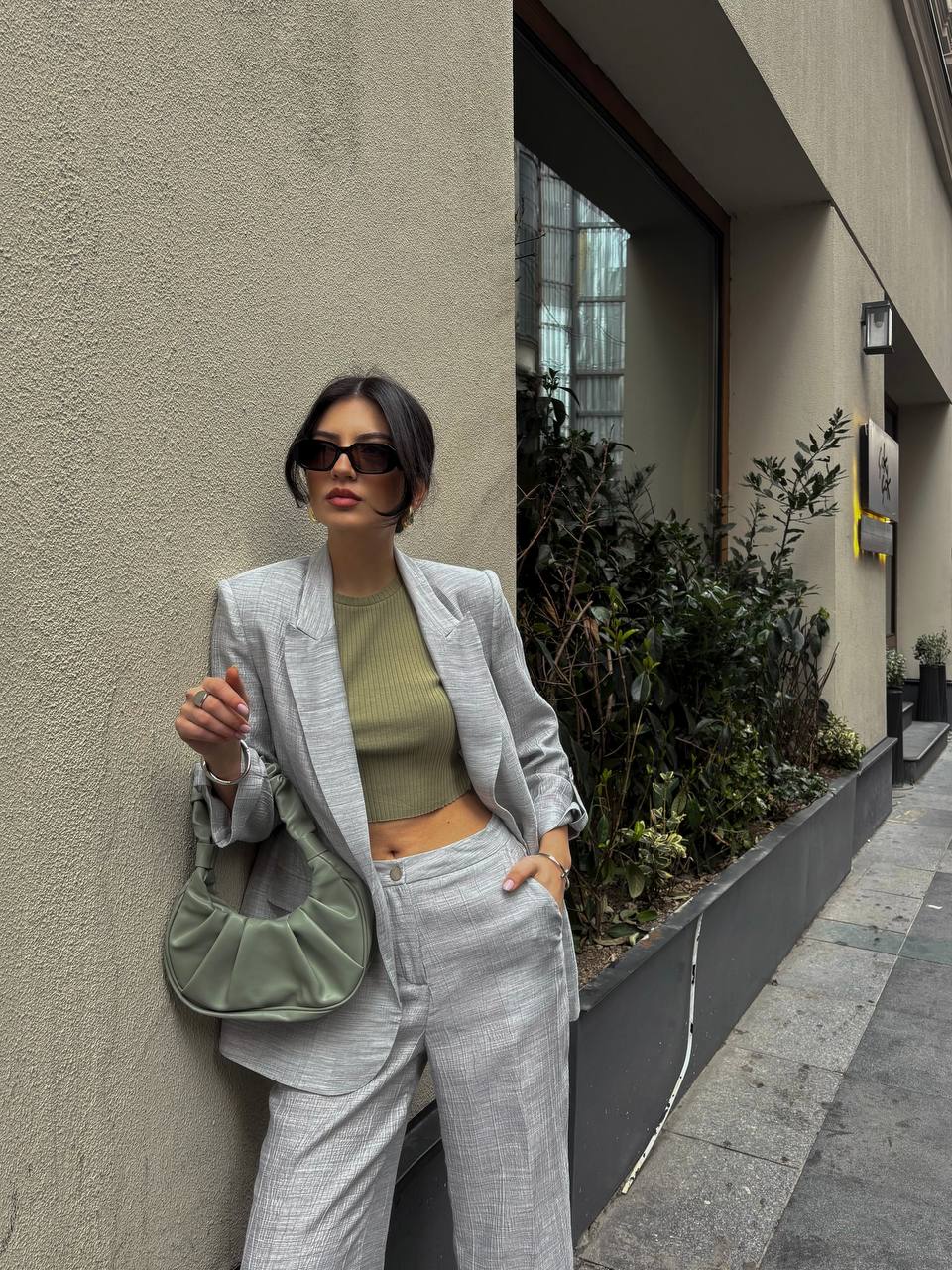 Woman in a gray suit with a green top and bag leaning against a wall.