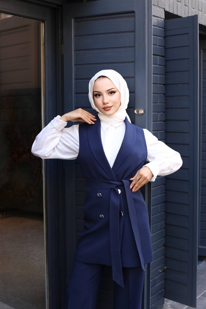 Woman wearing a navy blue vest and pants outfit with a white shirt, standing against a dark blue wall.