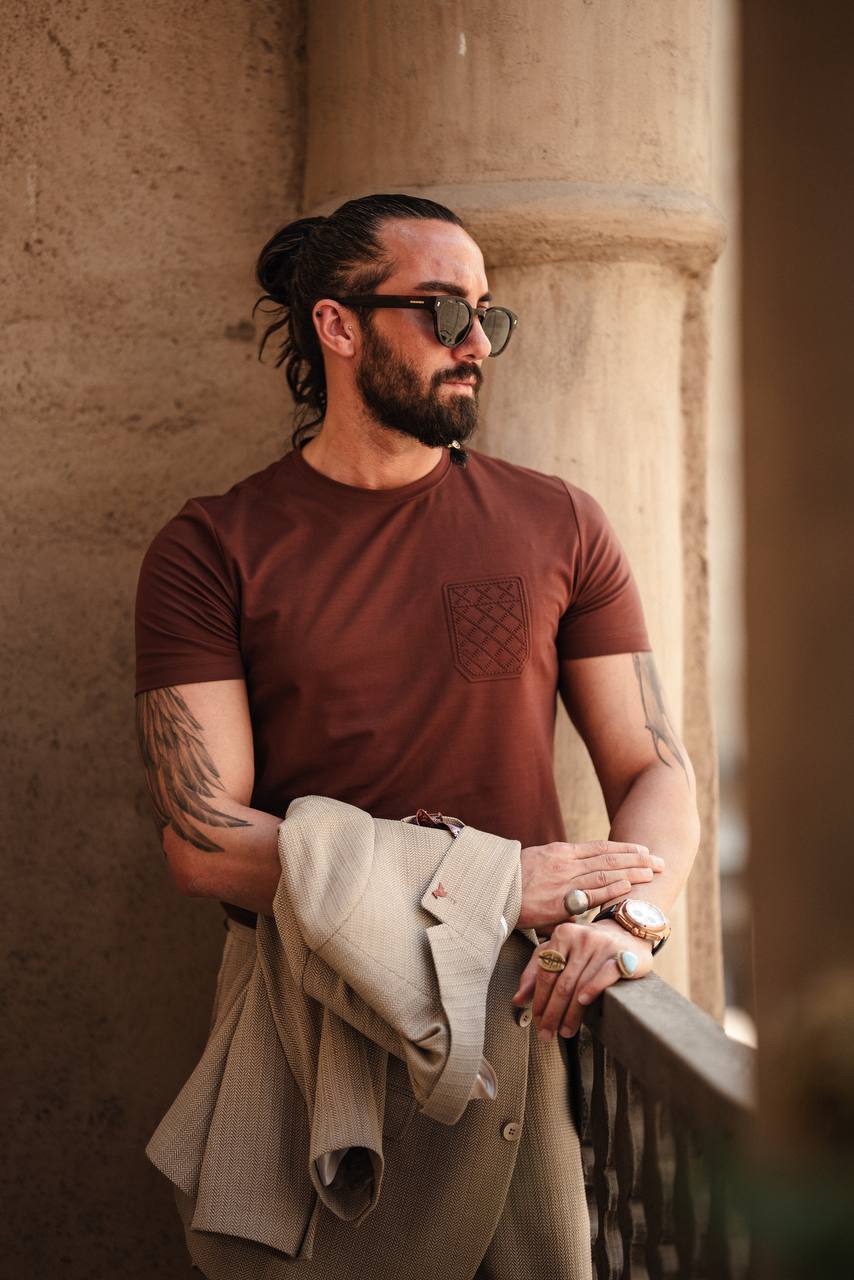 A man wearing a rich earth brown knit tee with a subtle chest pocket, paired with light-colored shorts, holding a jacket over his arm, standing in a casual pose.