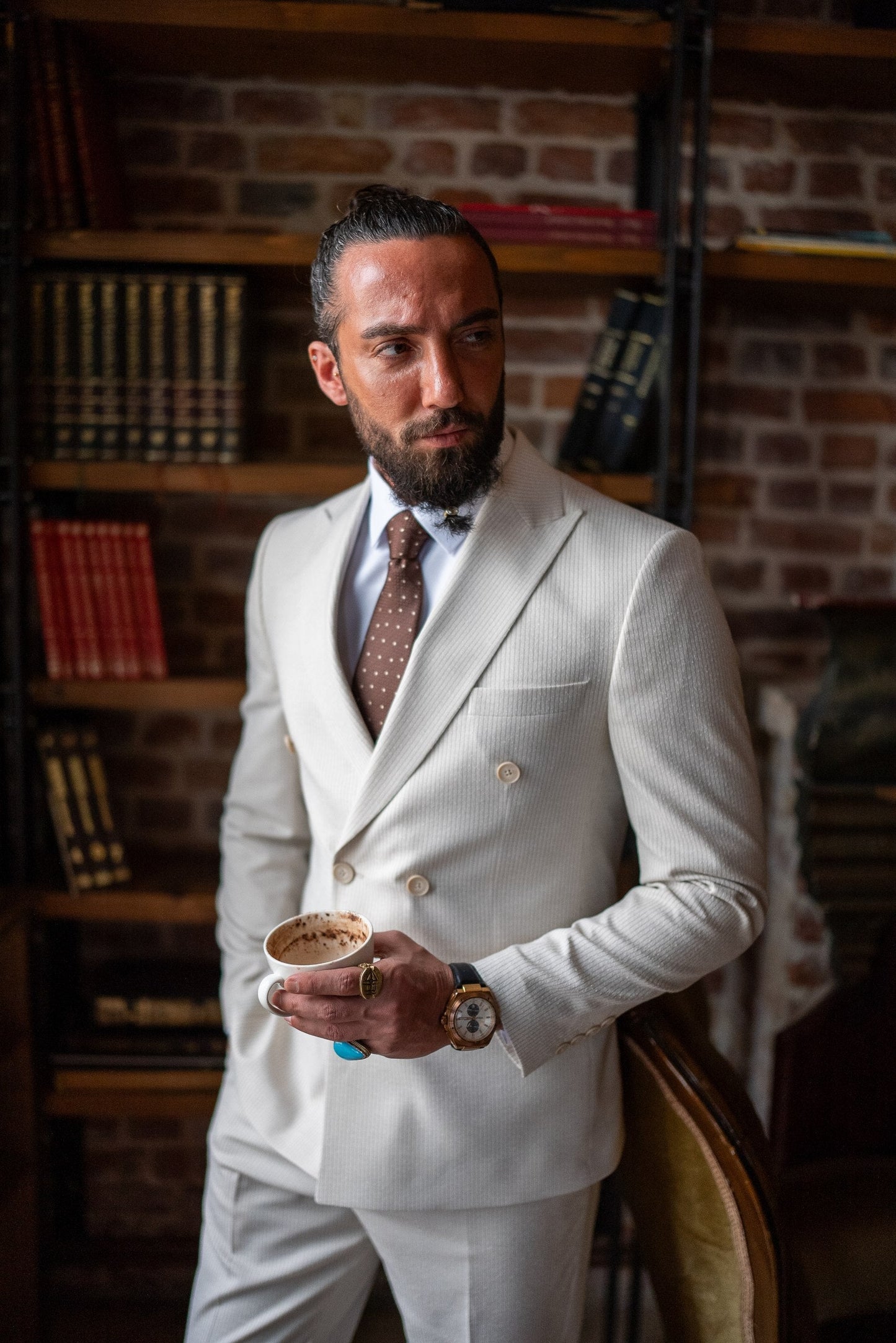 Man in a white suit holding a coffee cup in a room with brick walls and bookshelves.