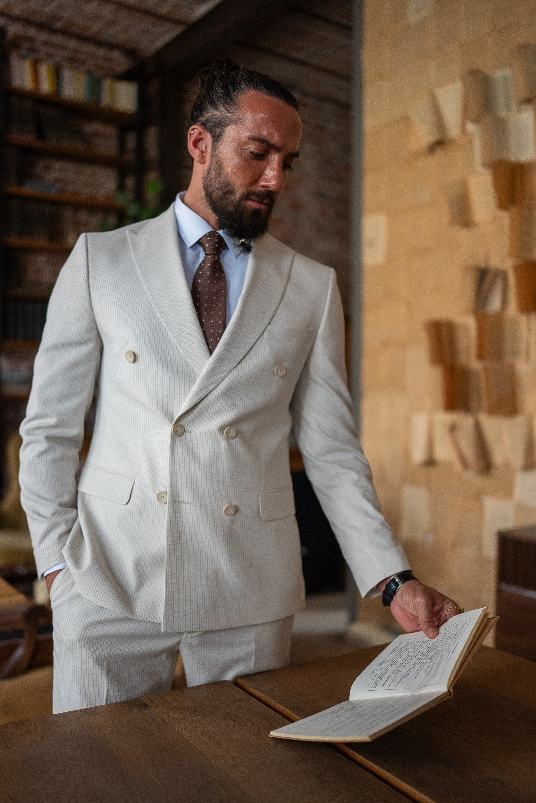 Man in a beige suit reading a book in a room with wooden walls and shelves.
